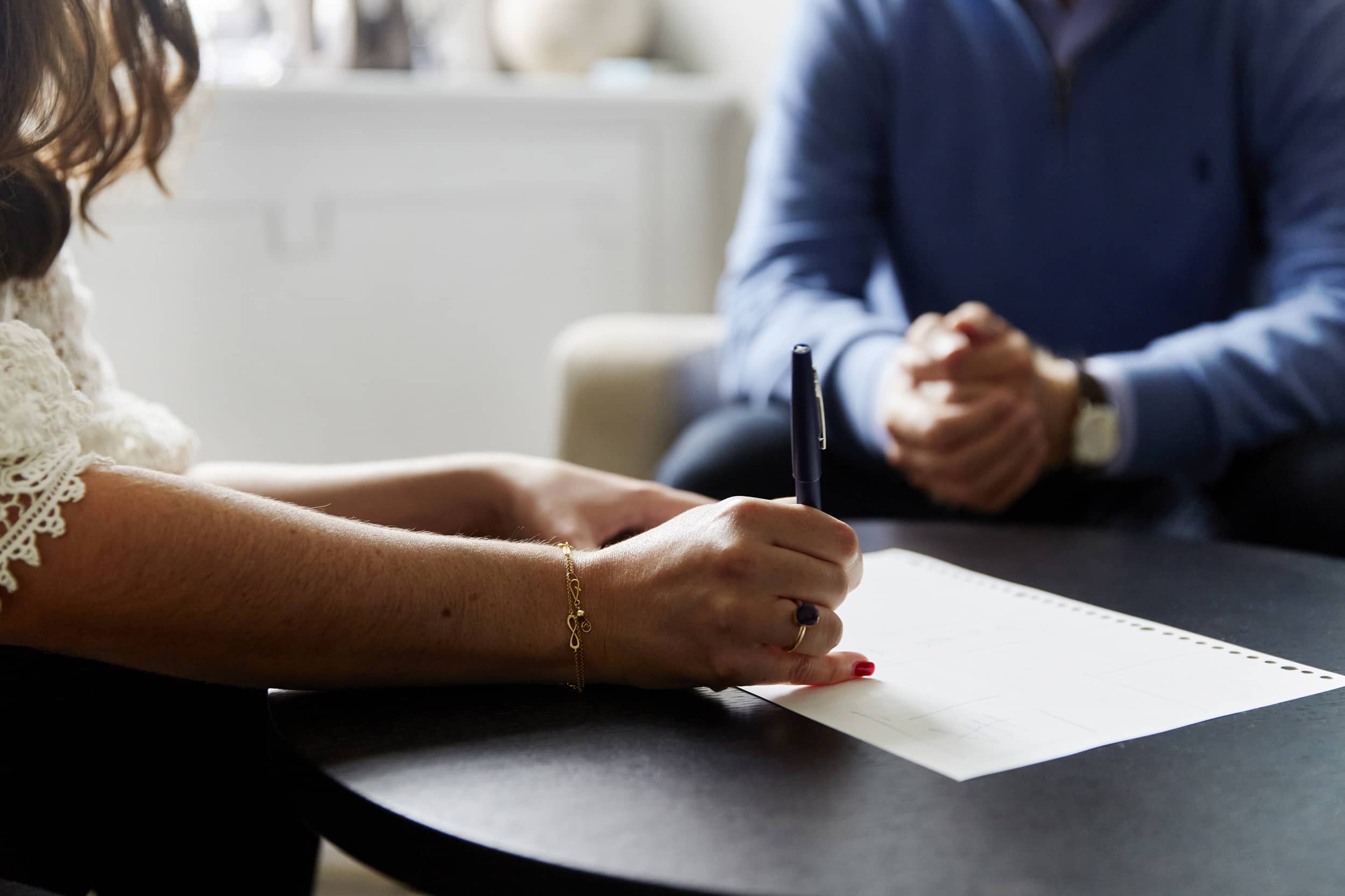 Close-up of a therapist taking notes on paper, with a client in a blue jumper seated in the background during a private consultation session at The Chelsea Psychology Clinic.