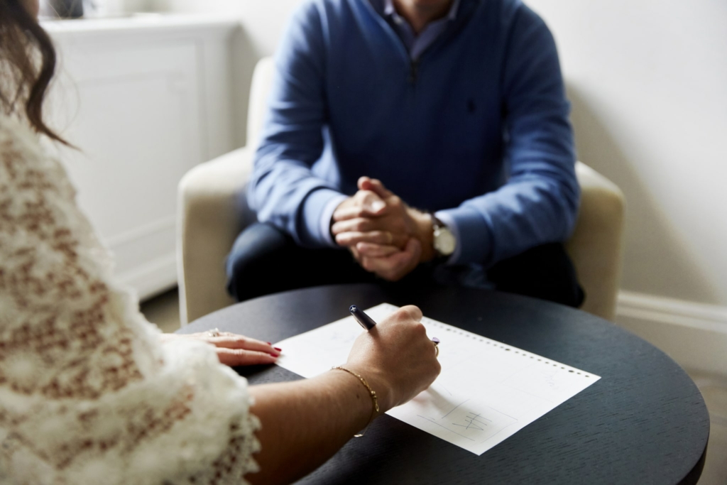 Close-up of a therapist taking notes on paper, with a client in a blue jumper seated in the background during a private consultation session at The Chelsea Psychology Clinic.