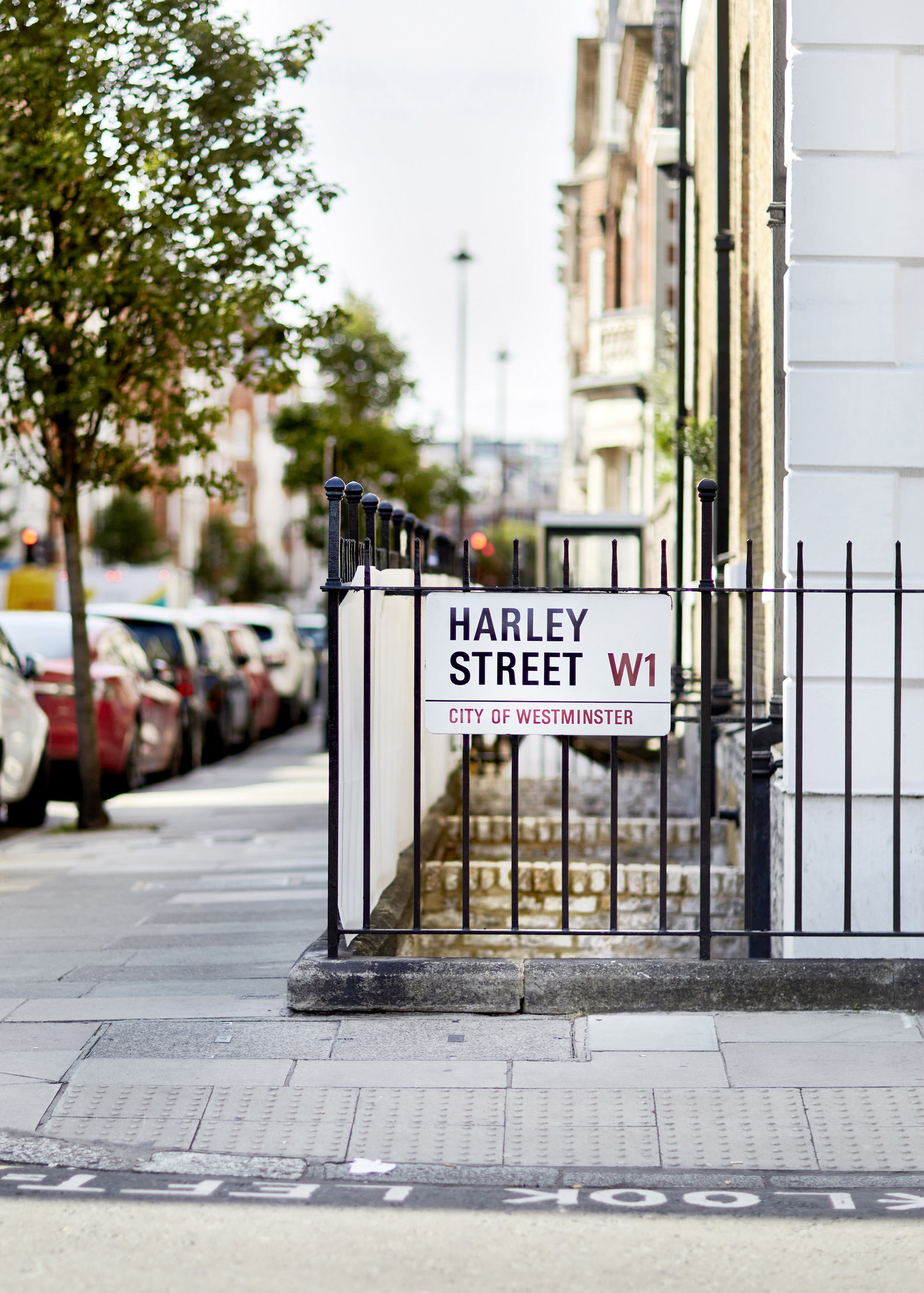 Harley Street W1 sign in the City of Westminster, London, renowned for its prestigious medical and psychological clinics, with a row of parked cars and historic buildings in the background.