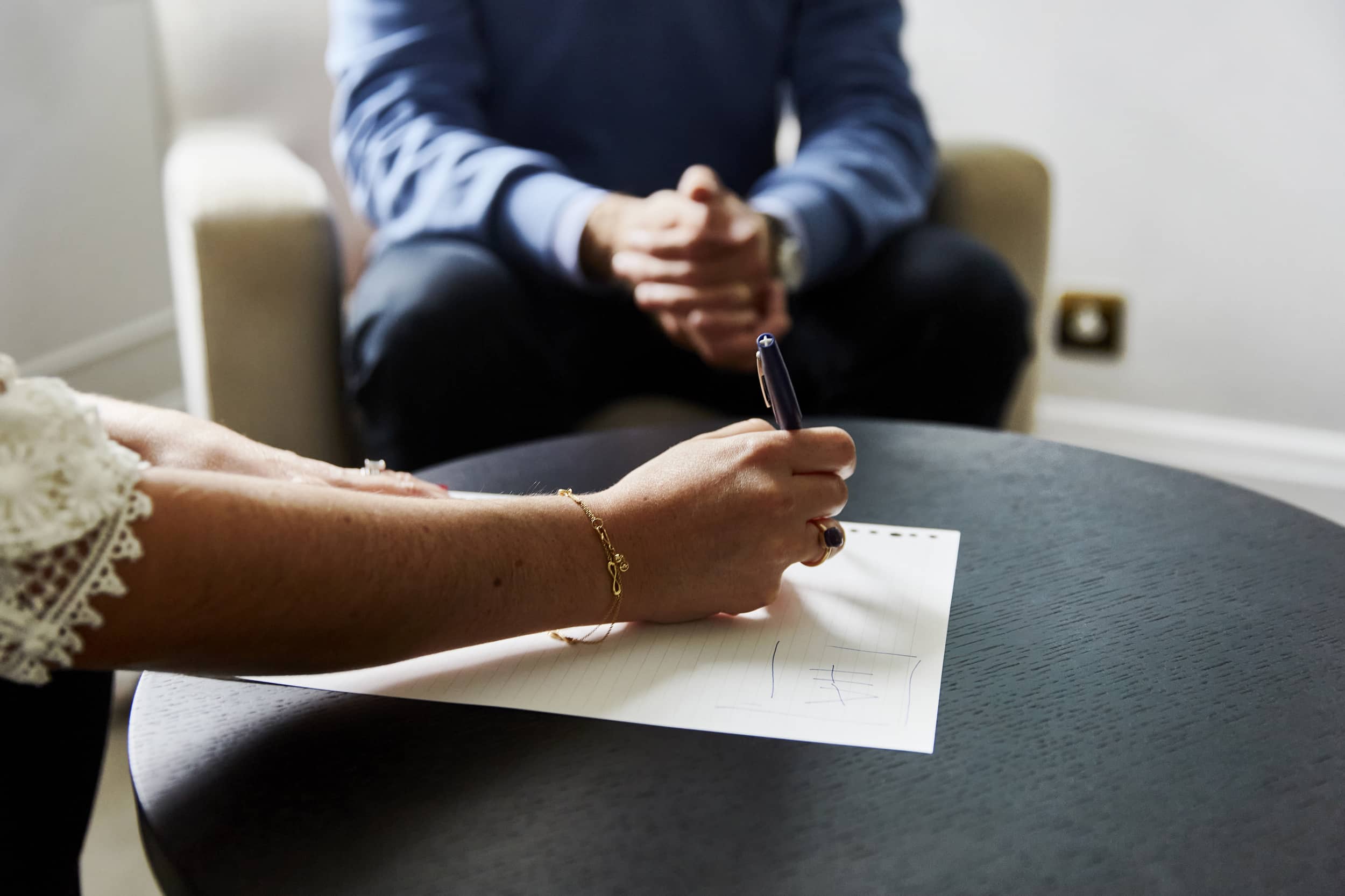 Close-up of a therapist taking notes on paper, with a client in a blue jumper seated in the background during a private consultation session at The Chelsea Psychology Clinic.
