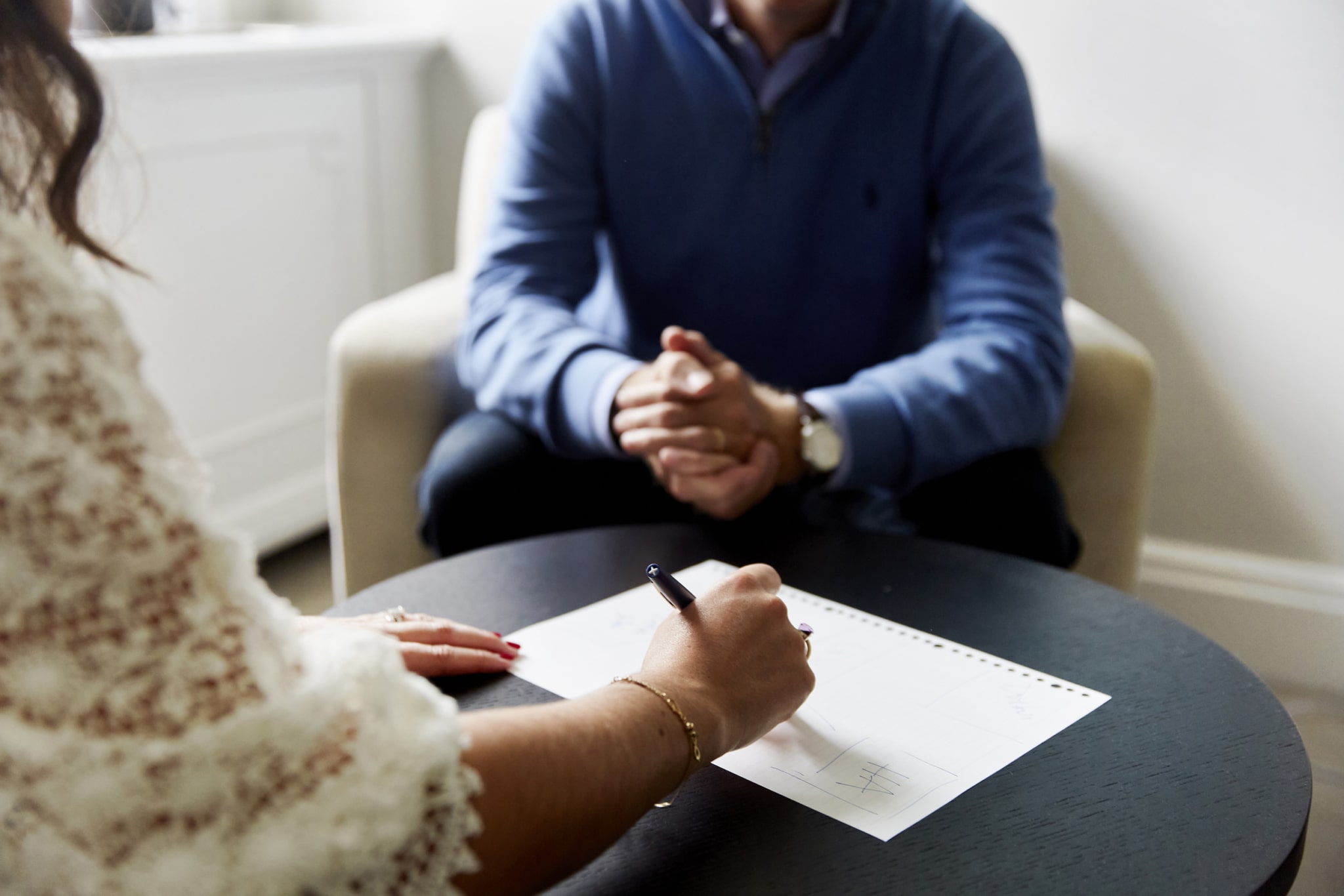 Close-up of a therapist taking notes on paper, with a client in a blue jumper seated in the background during a private consultation session at The Chelsea Psychology Clinic.