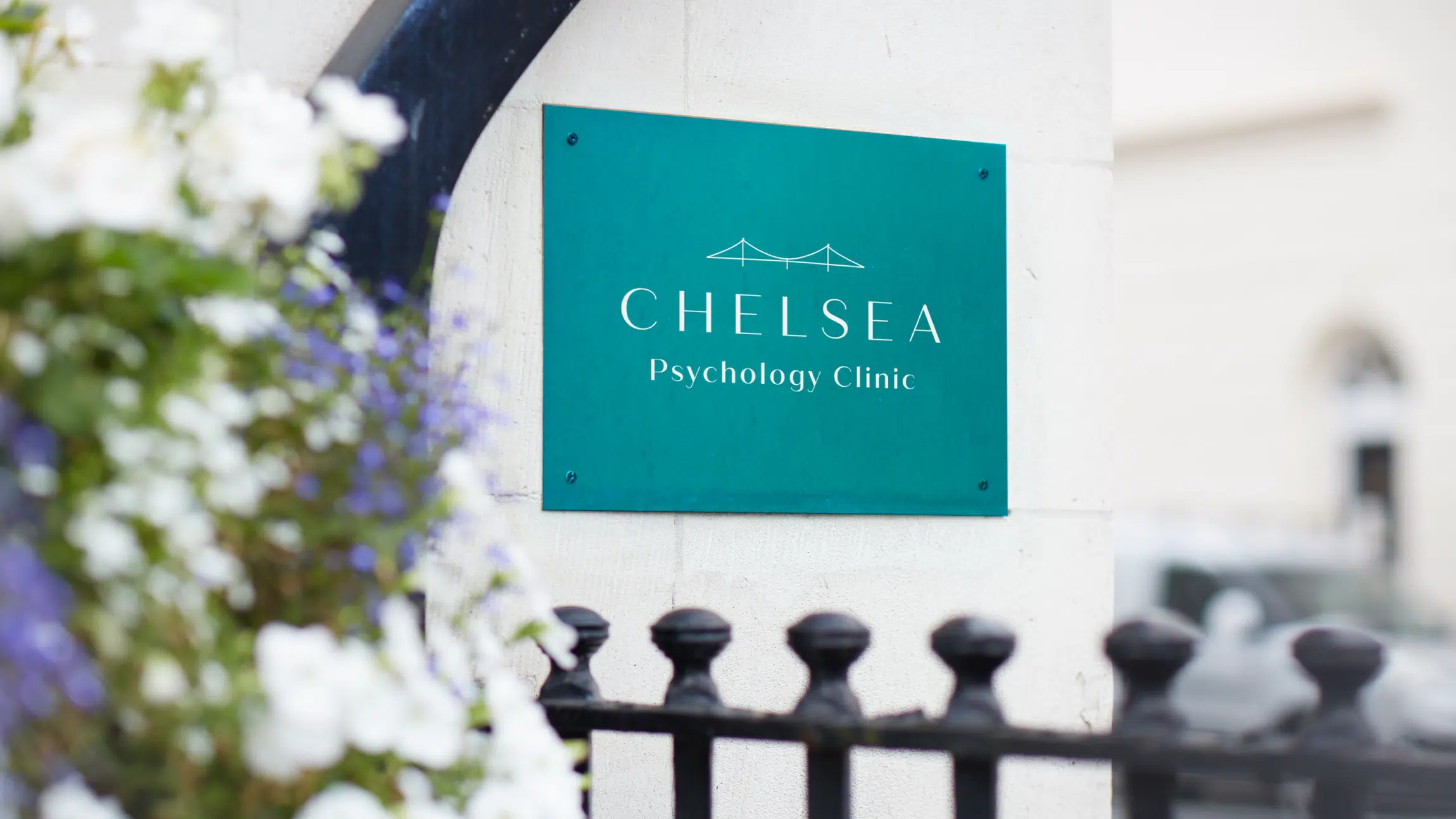 Green ‘Chelsea Psychology Clinic’ sign on a cream stone wall in London, with white and purple flowers in the foreground, representing a private therapy and psychiatry practice.